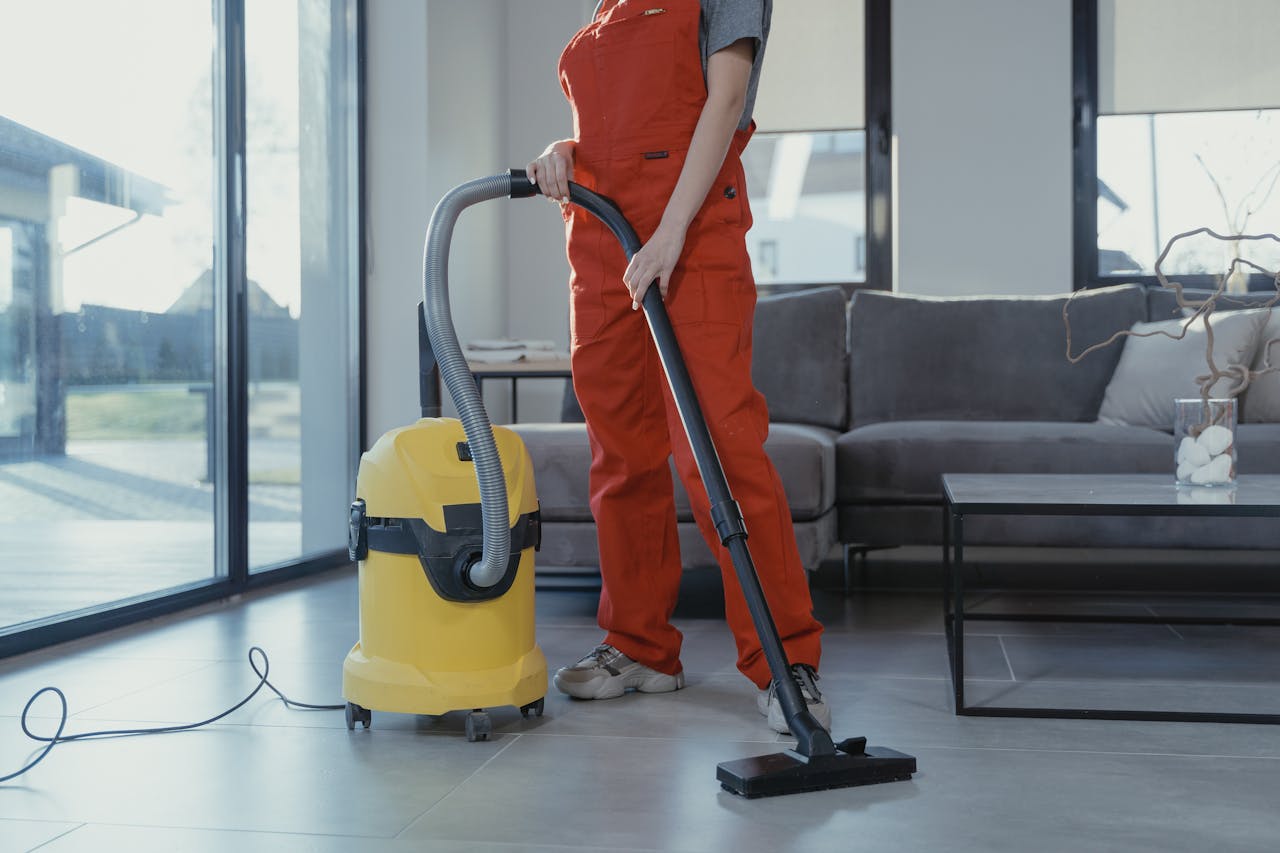 services-04 A professional cleaner in red coveralls vacuuming a modern living room with a yellow vacuum cleaner.