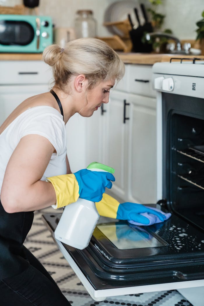 our-story A woman uses a spray bottle and cloth to clean an oven in a modern kitchen setting.
